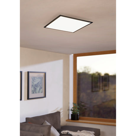 Frinarne square LED panel light, 59.5 x 59.5cm, with slim black aluminium frame and white diffuser, shown flush-mounted on a living room ceiling near a window and sofa.