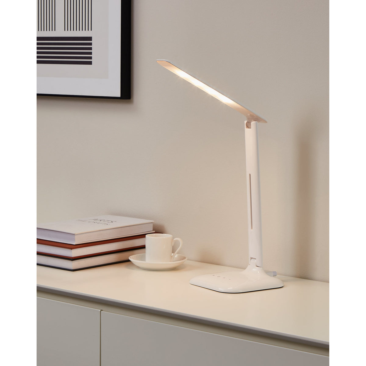 Frolmora LED touch table lamp in white, folding flat bar head on a slim steel and plastic stem with square base, shown lit on a sideboard beside books and a cup.