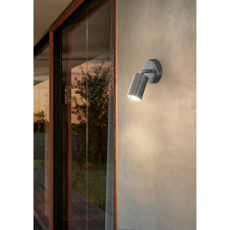 Frosmenne LED Outdoor Wall Light in stainless steel, mounted beside a glazed door with a round wall plate, adjustable arm and cylindrical head casting a warm downward glow at dusk.