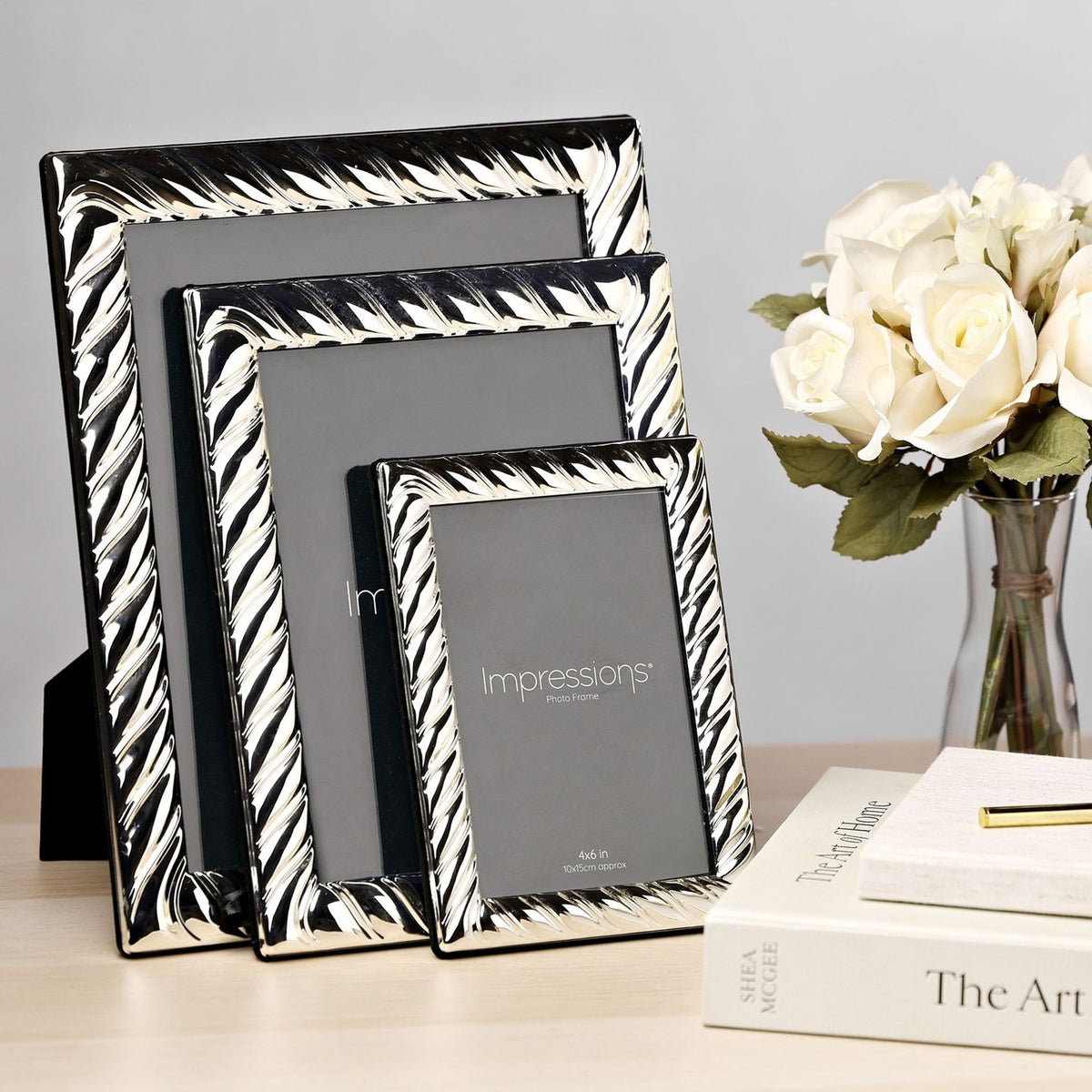 Three rectangular silverplated photo frames with swirl-textured borders and built-in stands, shown in graduating sizes on a desk beside books and white roses.