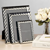 Three rectangular silverplated photo frames with swirl-textured borders and built-in stands, shown in graduating sizes on a desk beside books and white roses.