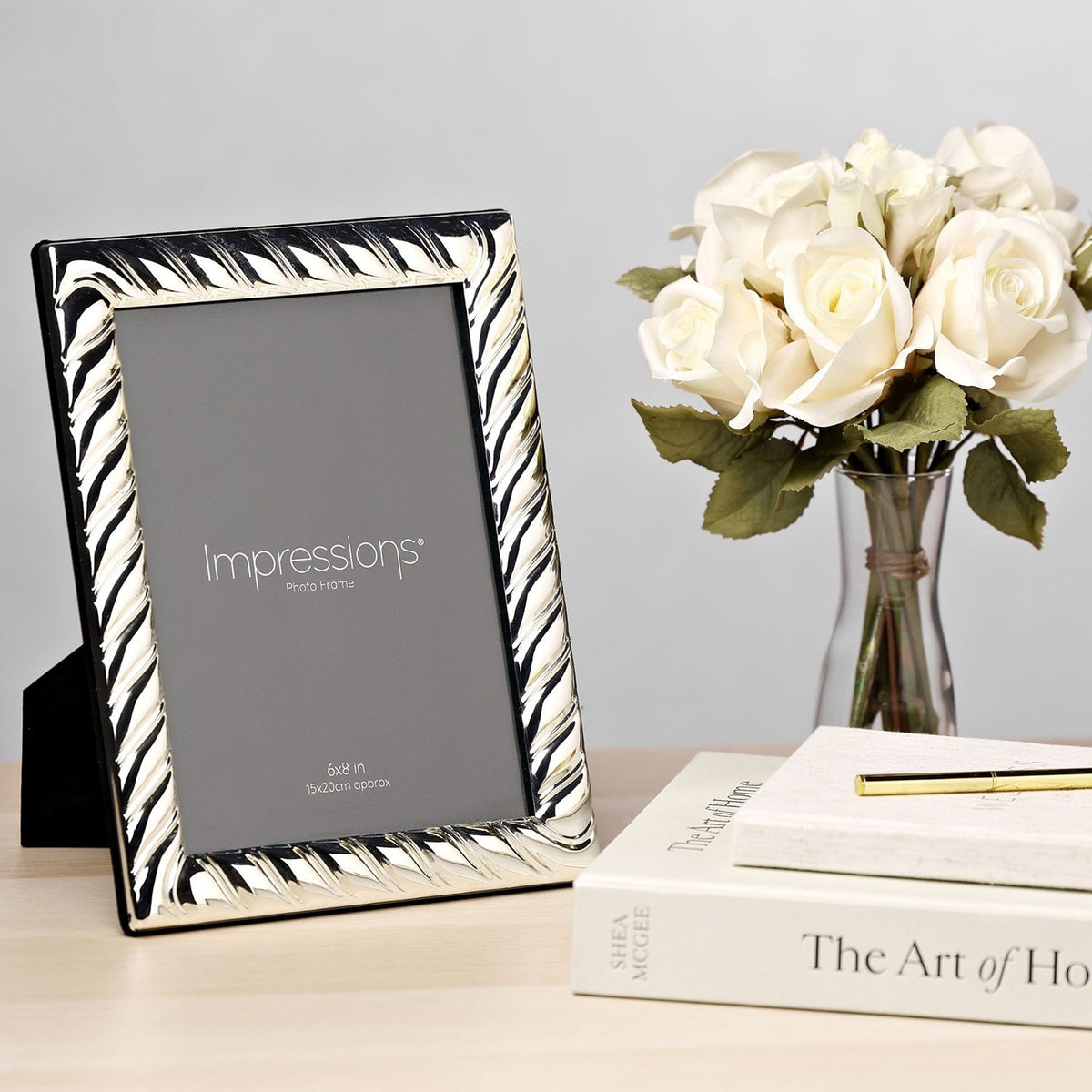 Rectangular silverplated photo frame with diagonal swirl textured border and black stand, shown on a desk beside white roses and books.