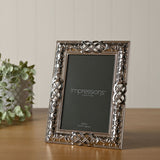 Rectangular silver metal photo frame with Celtic knot and scrollwork detailing, holding a 4x6in placeholder, displayed upright on a wooden surface with a potted plant nearby.