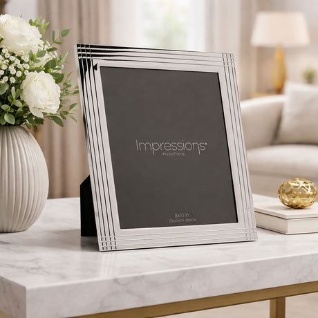 Rectangular silver plated metal photo frame with grooved striped border, holding a 10 x 8in portrait print, displayed on a marble table beside a vase of white flowers.