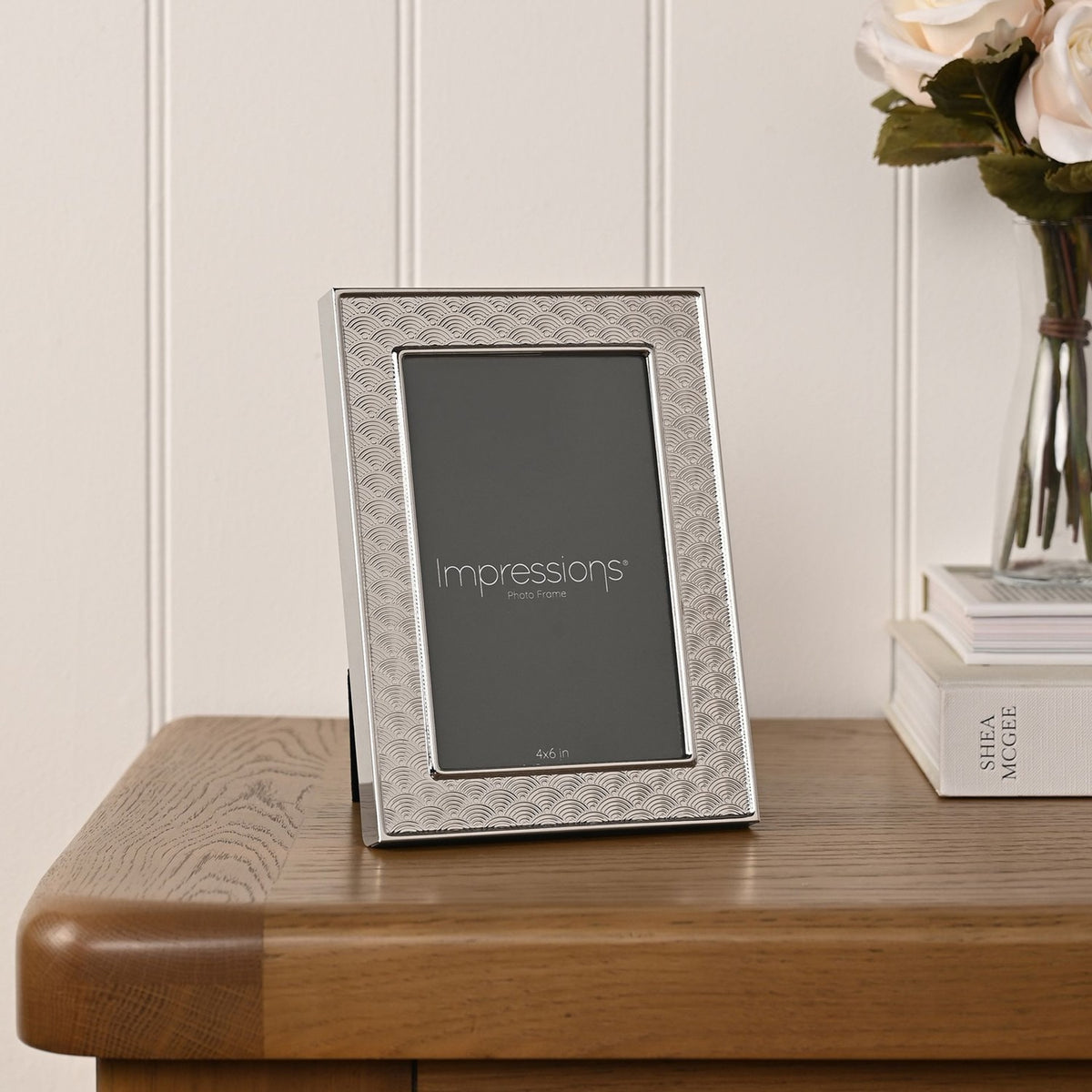 Art Deco Scallop silverplated metal photo frame with raised inner rim, shown upright on a wooden table beside books and a vase of flowers.