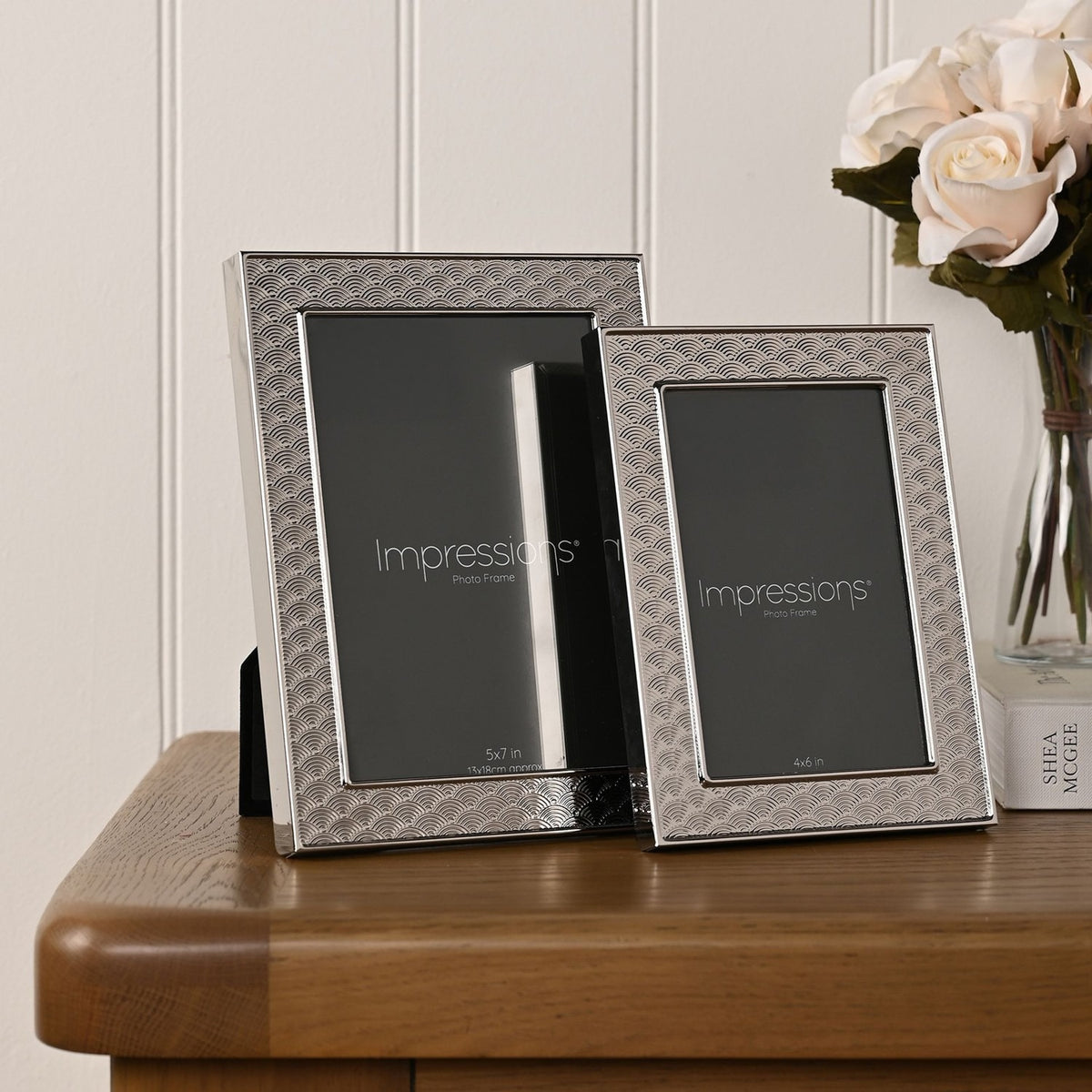 Two silverplated Art Deco scallop-pattern photo frames, 5x7in and 4x6in, displayed on a wooden table beside white roses.
