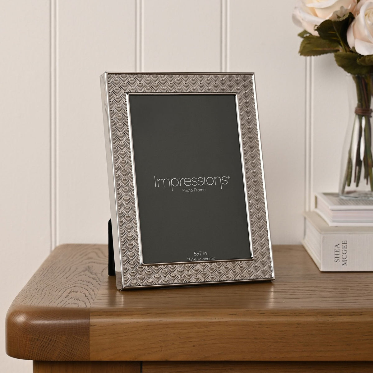 Rectangular silverplated photo frame with art deco scallop pattern border, displayed upright on a wooden table beside books and flowers.