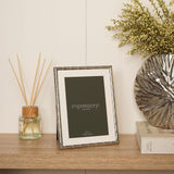 Silver metal photo frame with criss cross pattern and white mount, 5 x 7in, displayed upright on a wooden sideboard beside a reed diffuser and vase.