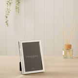 Narrow rectangular silverplated metal photo frame with a slim polished border, shown upright on a wooden surface beside a reed diffuser in a living room setting.