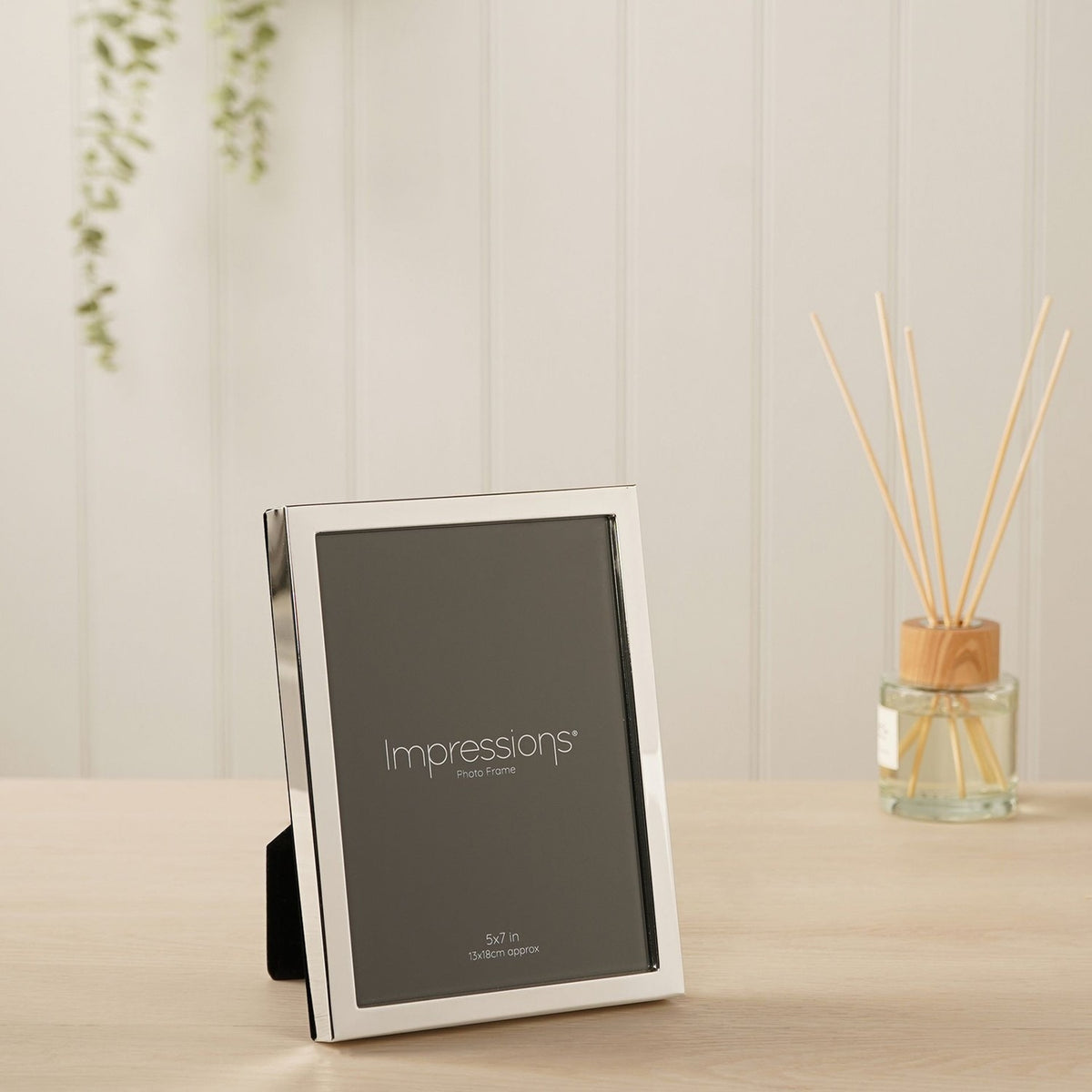 Narrow rectangular silver-plated metal photo frame, 5x7in, with a slim polished silver border, shown upright on a wooden surface in a living room setting.