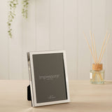 Narrow rectangular silver-plated metal photo frame, 5x7in, with a slim polished silver border, shown upright on a wooden surface in a living room setting.