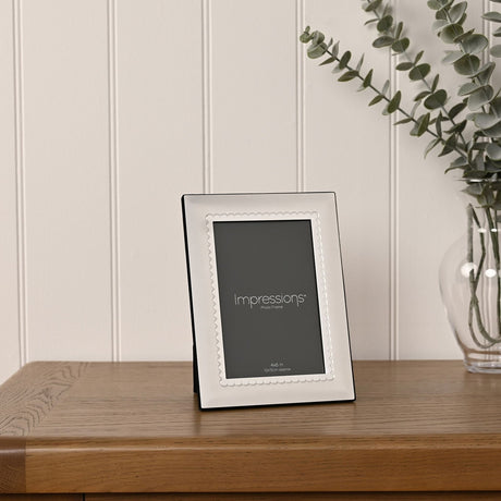 Scalloped freestanding photo frame in satin silver plated metal with a rectangular opening, displayed upright on a wooden sideboard beside a vase of eucalyptus leaves.