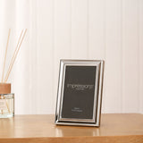 Moulded freestanding rectangular photo frame in silverplated metal, shown on a wooden table with a reed diffuser against a light panelled wall.