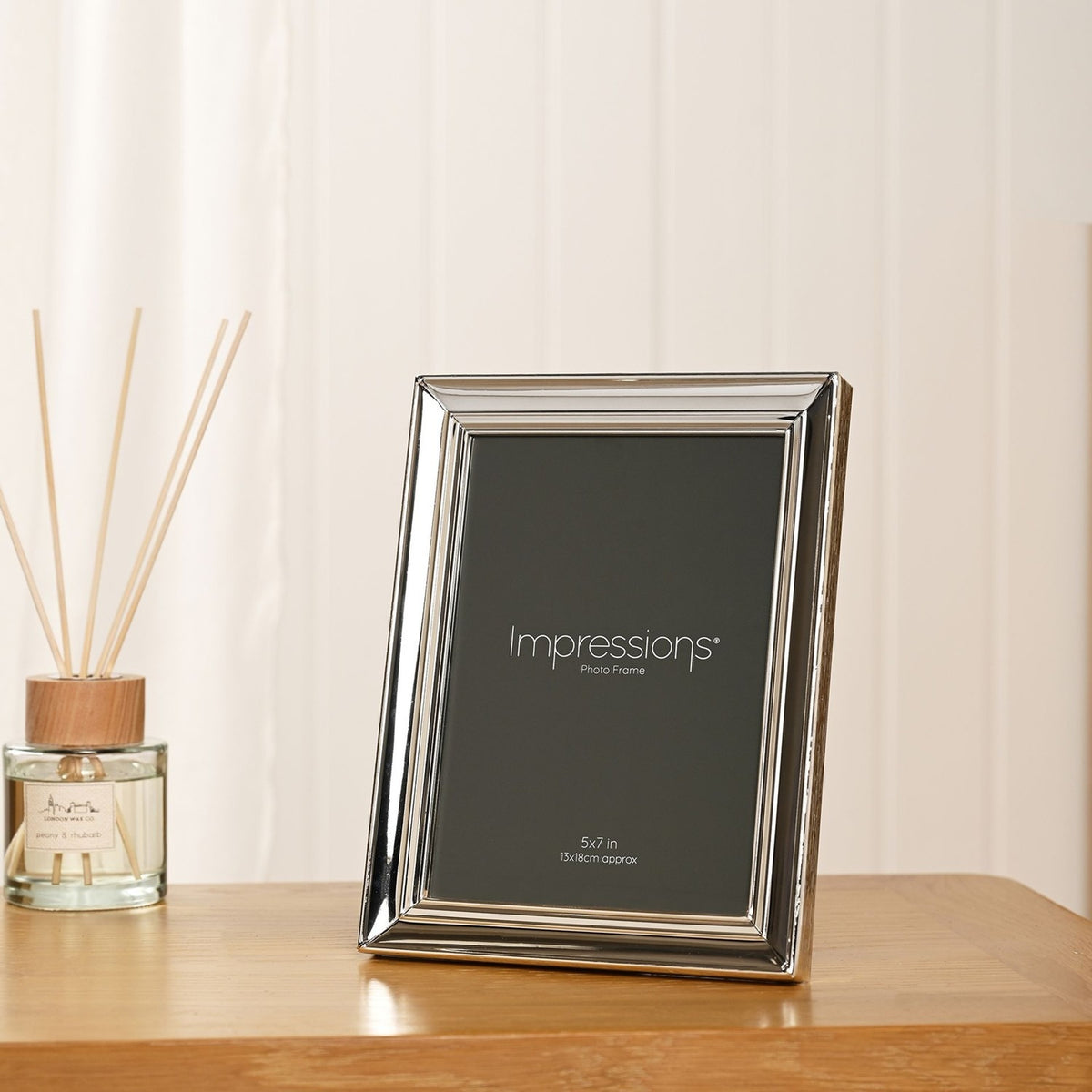 Rectangular freestanding photo frame in moulded silverplated metal with tiered profile, shown on a wooden table beside a reed diffuser, holds a 5x7in print.
