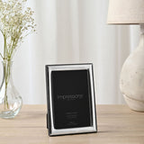 Silver-plated narrow rectangular photo frame with smooth shiny border and wooden back, holding a 6 x 4in print, displayed on a table beside a vase and lamp.