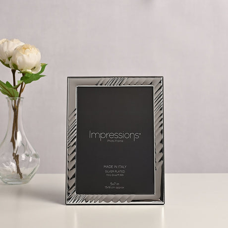 Rectangular silver-plated photo frame with textured diagonal wave border and smooth inner edge, holding a 7 x 5in print, displayed beside white flowers on a light background.