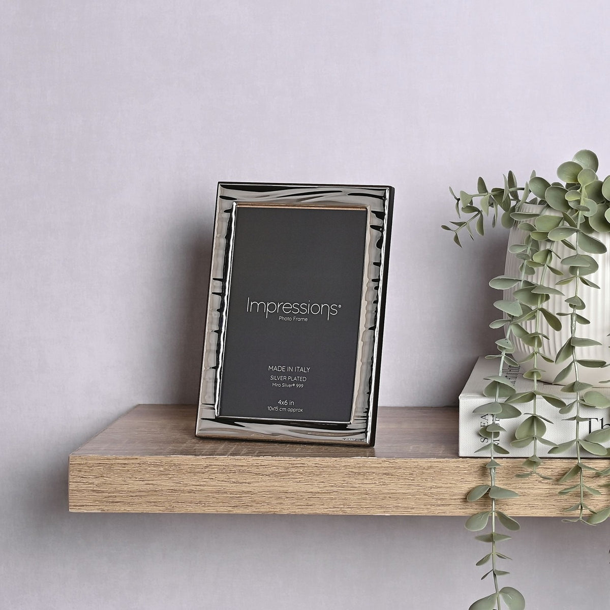 Italian Narrow Wave Photo Frame 6 x 4in in silver with rippled wave-textured border, displayed upright on a wooden shelf beside books and trailing greenery.