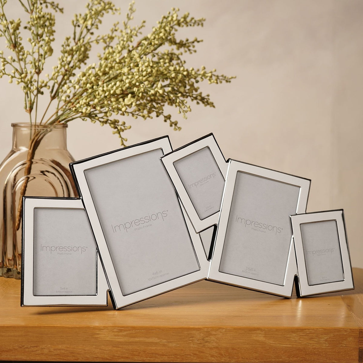 Overlapping multi-aperture photo frame in silver-plated metal with five tilted rectangular openings, shown on a wooden surface beside a vase of dried foliage.
