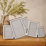 Overlapping multi-aperture photo frame in silver-plated metal with five tilted rectangular openings, shown on a wooden surface beside a vase of dried foliage.