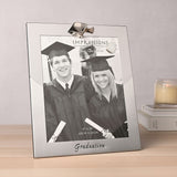 Silver two-tone metal photo frame for 10 x 8in photo, with a mortarboard cap detail on top and "Graduation" script at the base, shown on a wooden surface.