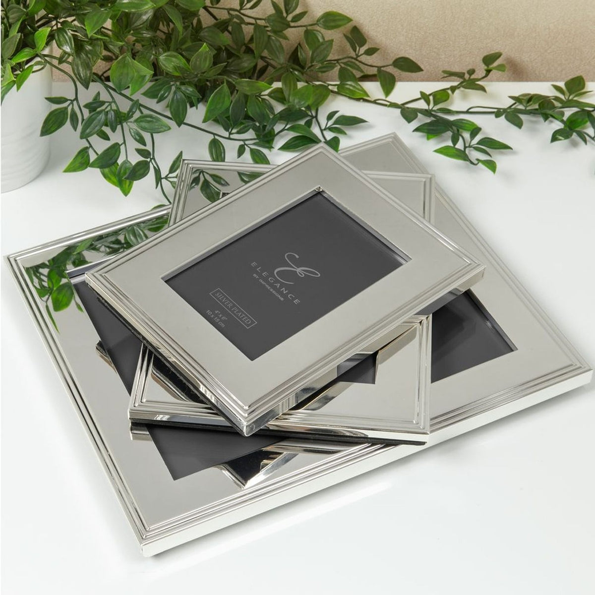 Stack of silverplated rectangular photo frames with ribbed-edge borders in silver metal finish, displayed on a white surface with greenery in the background.