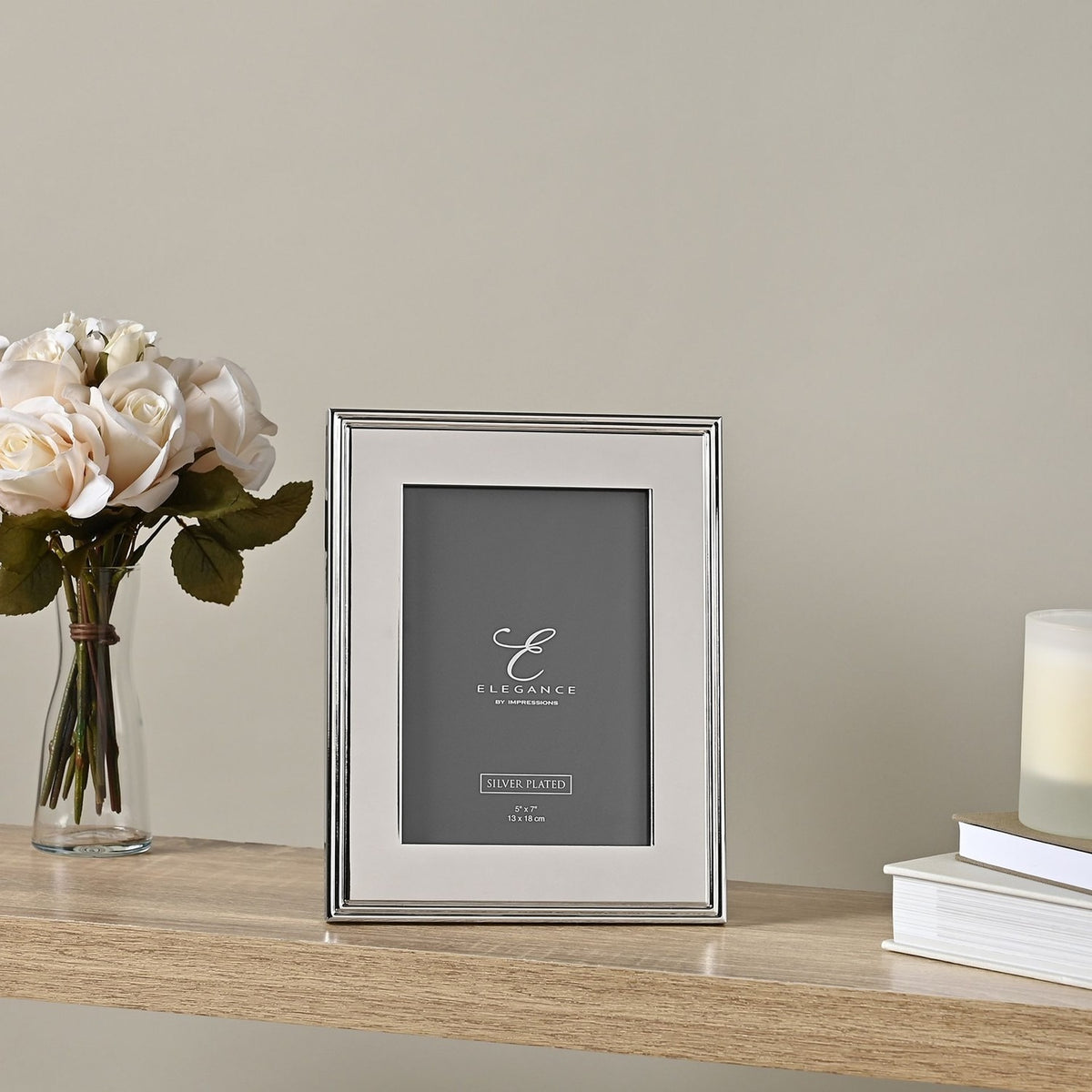 Rectangular silverplated metal photo frame with ribbed edge border, displayed upright on a wooden shelf beside white roses and a candle.