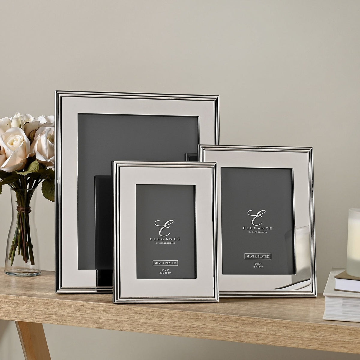 Three silverplated rectangular photo frames in graduated sizes with ribbed-edge borders, shown on a wooden table beside roses and books.