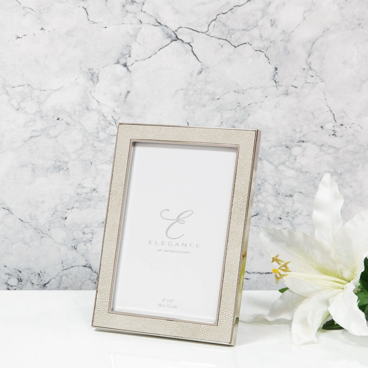 Rectangular photo frame in cream faux shagreen with a silver-tone metal edge and rear strut, shown against a marble background with a white lily.