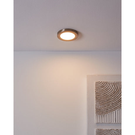 Fueva Flex LED downlight recessed into a ceiling with satin nickel rim and white diffuser, casting warm white light onto a plain wall below.