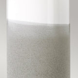 Close-up image of a Fulwell 1 Light Table Lamp in White Gloss and Matt Grey, featuring a glass container filled with a light gray liquid. The liquid's surface is akin to a delicate emulsion with tiny bubbles, resembling the lamp's modern textured design. The neutral and plain background enhances its contemporary feel.