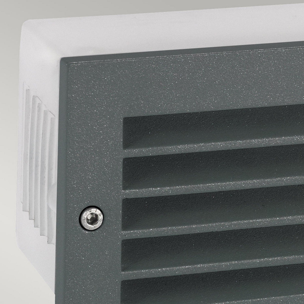 A close-up view of the Grimstad LED Recessed Wall Light - Graphite features a gray, vented surface with horizontal slats and a visible screw on the side. The graphite finish enhances its sleek appearance against the light gray background while offering energy-efficient illumination from the side profile.
