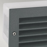 A close-up view of the Grimstad LED Recessed Wall Light - Graphite features a gray, vented surface with horizontal slats and a visible screw on the side. The graphite finish enhances its sleek appearance against the light gray background while offering energy-efficient illumination from the side profile.
