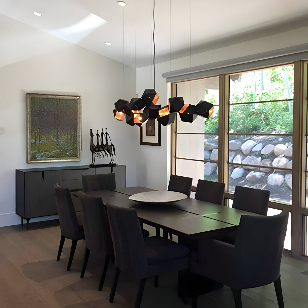 A modern dining room features a long dark wooden table surrounded by eight black upholstered chairs. The room is beautifully illuminated by the Spar 3 Light Pendant in Black & Gold, which adds a stylish touch to the contemporary lighting. Large windows bring in natural light, and a sideboard along with artwork decorates the white walls.