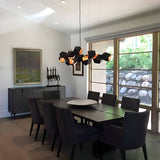 A modern dining room features a long dark wooden table surrounded by eight black upholstered chairs. The room is beautifully illuminated by the Spar 3 Light Pendant in Black & Gold, which adds a stylish touch to the contemporary lighting. Large windows bring in natural light, and a sideboard along with artwork decorates the white walls.