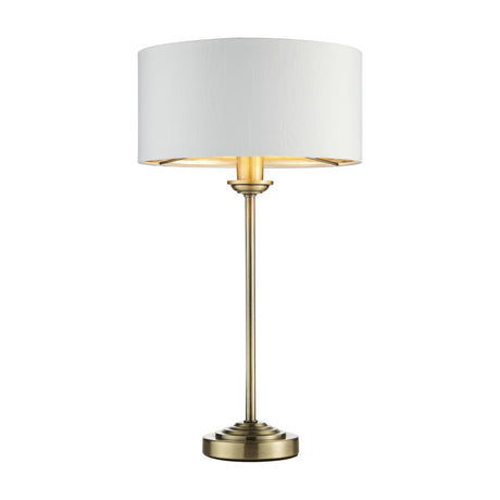 Gallery Direct Clervorne Table Lamp with slim antique brass stem, stepped base and white fabric drum shade, shown illuminated on a plain background.