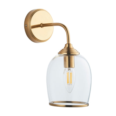 Gallery Direct Vellamis wall light with brushed aged brass arm, circular backplate and clear glass shade over a filament bulb, on a white background.