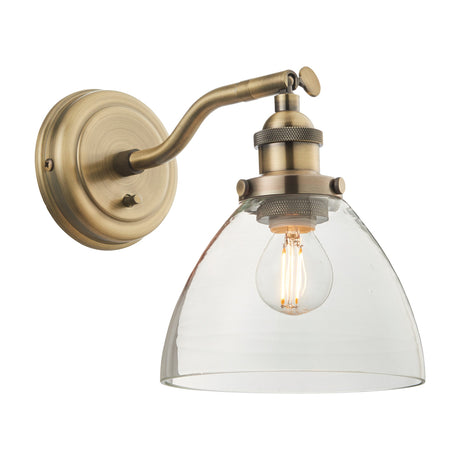 Sarwen Wall Light by Gallery Direct with antique brass adjustable arm, rocker switch backplate, and clear glass dome shade housing a filament bulb.