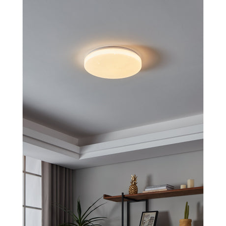 Galmane 38cm round LED flush ceiling light in white, illuminated with a crystal-effect diffuser, shown mounted on a light grey ceiling in a living room with a shelf below.