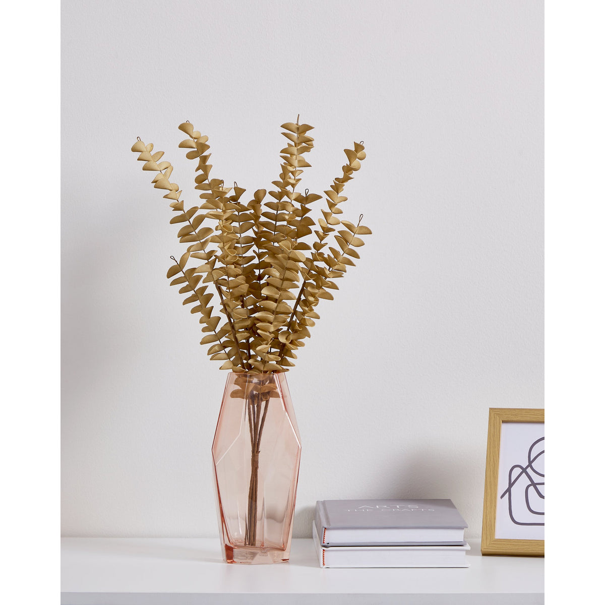 Gannan Artificial Leaf Spray in brown, tall faceted pink glass vase on a white shelf with stacked books and a framed print against a light grey wall.