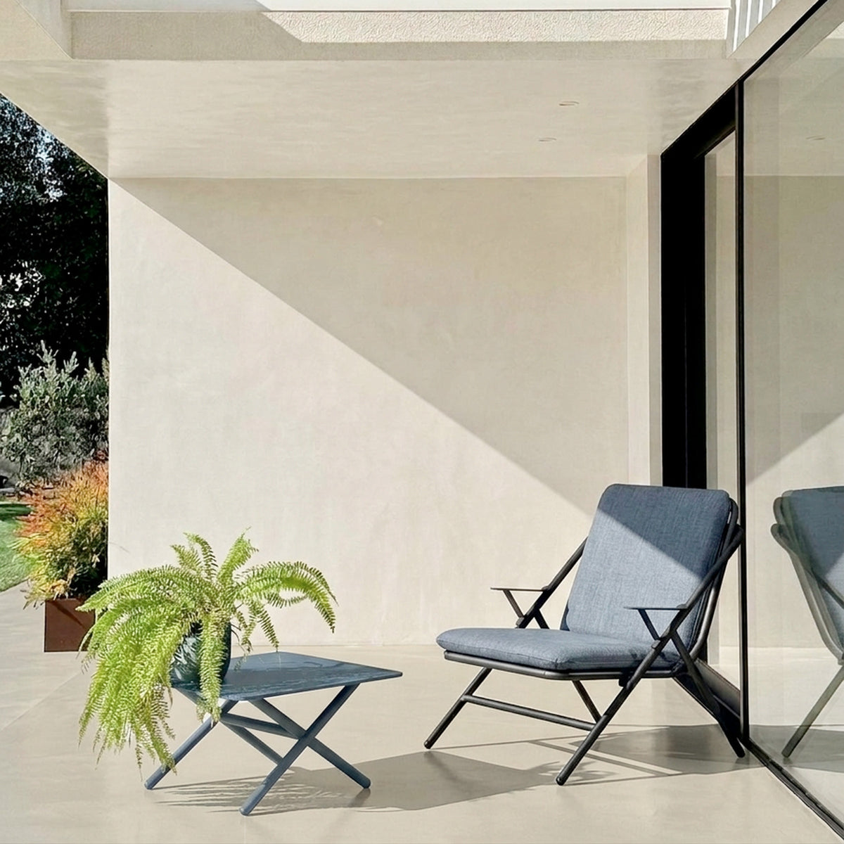 Garda square aluminium outdoor coffee table in graphite with X-shaped crossed legs, shown on a patio beside a matching lounge chair with blue cushions.