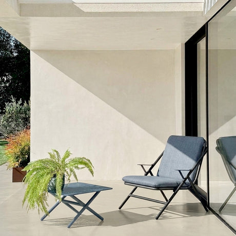 Garda square aluminium outdoor coffee table in graphite with X-shaped crossed legs, shown on a patio beside a matching lounge chair with blue cushions.