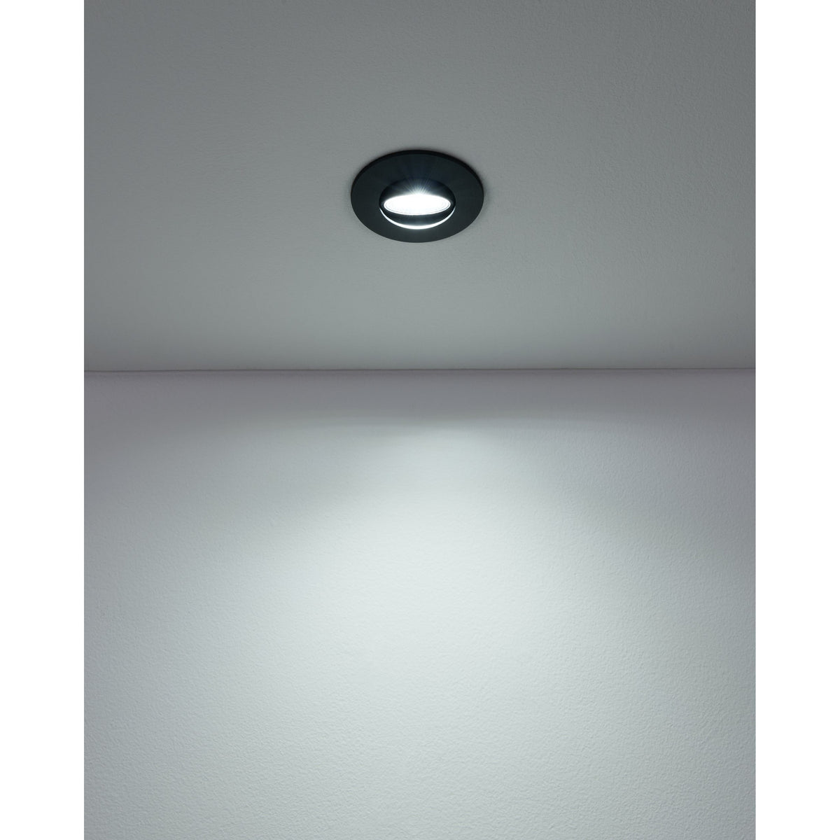 Gedrez round LED downlight recessed in a plain ceiling, black finish, casting a bright cool-white light downward onto a grey wall and floor.