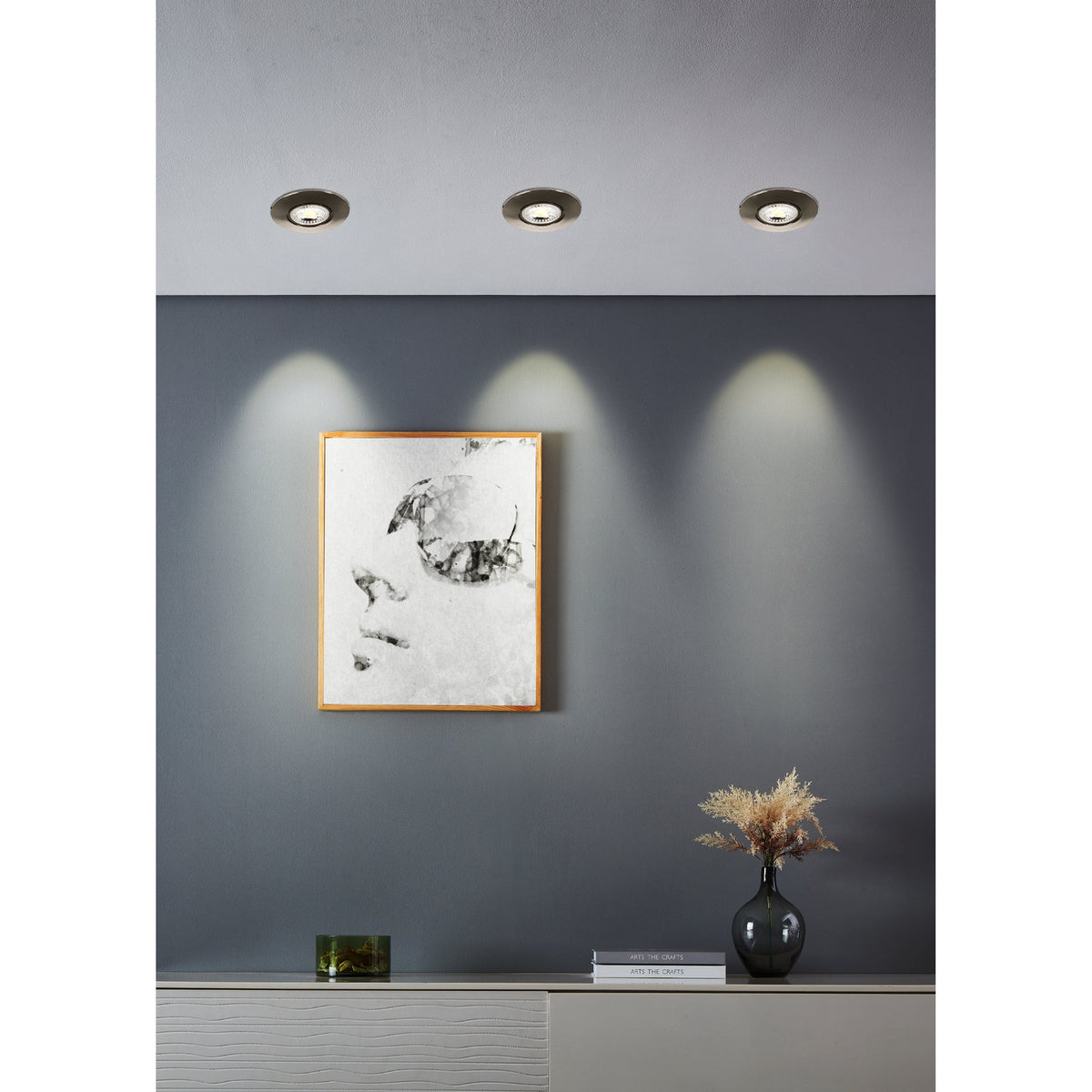 Three round Gedrez LED downlights recessed in a ceiling with satin nickel finish, casting warm light onto a grey wall above a framed portrait print.