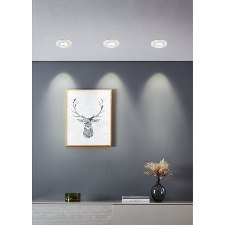 Three round white LED downlights recessed in a ceiling, casting warm light down a grey wall above a framed stag print and sideboard with vase and books.