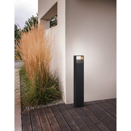 Gelbora LED bollard light in anthracite, a slim aluminium post with a rectangular top, shown lit on a wooden deck beside a house wall and ornamental grasses.