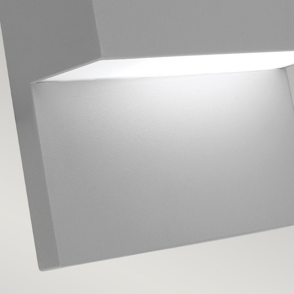 Close-up of the Geneve Outdoor 1 Light Wall Lantern in silver, featuring a modern, square-shaped design crafted from die-cast aluminium. The lantern is slightly angled, highlighting its smooth surface and bright white illumination emanating from within. It exudes elegance against a soft gray background, making it ideal for outdoor lighting.
