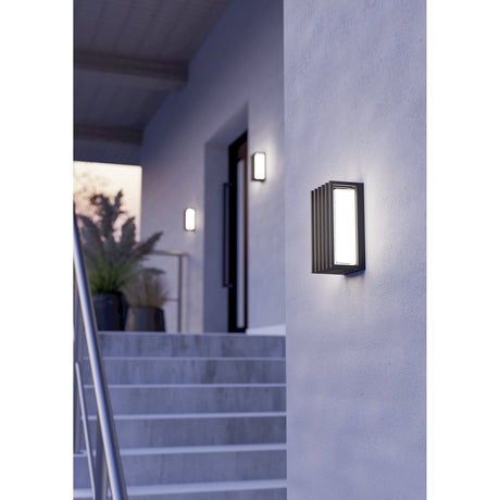 Genovese LED outdoor wall light in black aluminium with stepped vertical slats and a clear rectangular shade, shown illuminated beside a front door on a light grey exterior wall.