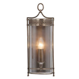 The Guildhall 1 Light Wall Light in dark bronze showcases a Georgian-inspired design with a vintage lantern shape. It features a clear cylindrical glass cover that encloses a single bulb, complemented by decorative metal accents and an easy-to-use hanging loop at the top.