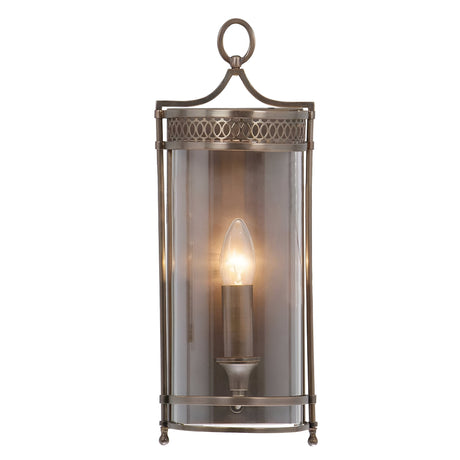 The Guildhall 1 Light Wall Light in dark bronze showcases a Georgian-inspired design with a vintage lantern shape. It features a clear cylindrical glass cover that encloses a single bulb, complemented by decorative metal accents and an easy-to-use hanging loop at the top.