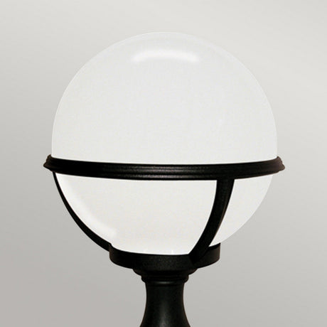 A Glenbeigh 1 Light Pedestal/Porch Lantern, featuring a round, white globe lamp within a black metal frame on a stand, evokes the essence of a modern outdoor lantern against a light gray background.